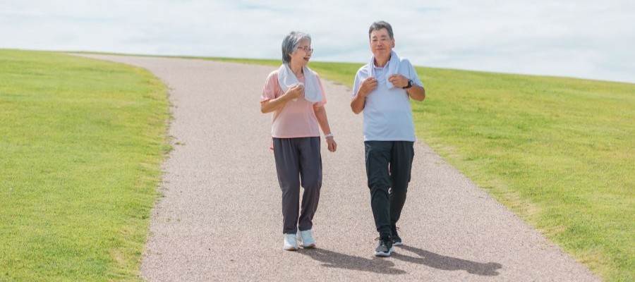 An older woman and man are out walking during the day. They are wearing comfortable clothes, sneakers and both have small towels around their necks.