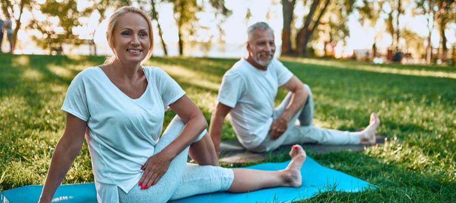 A woman and man sit on yoga mats in the grass with morning light coming through the trees. They are seated in a stretching position.