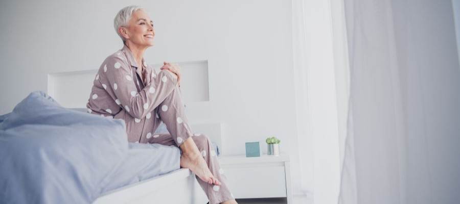 Short, white haired woman sits on the edge of the bed with a smile on her face.