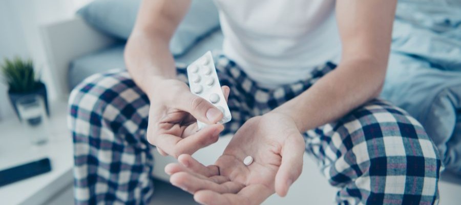 A person wearing blue checkered pajama pants sits on the edge of a bed with a medicine package in his hands.