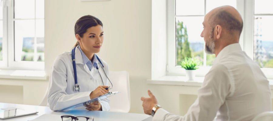 A man sits across from a doctor wearing a lab coat with a stethoscope around her neck and a clipboard in her hand.