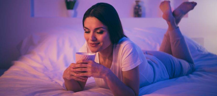 A woman is lounging on her stomach, on a bed, holding cup in her hands.