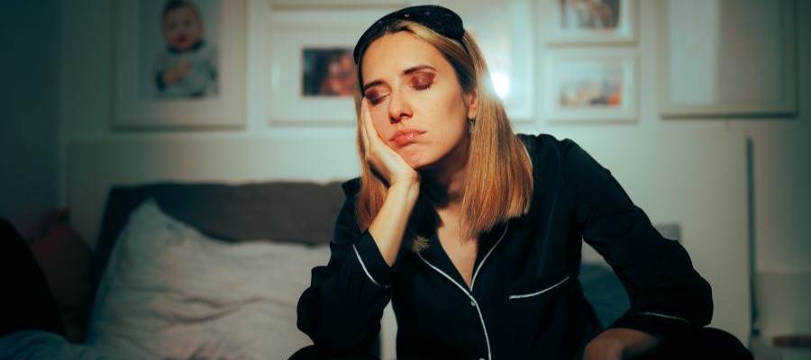 A woman sits on a bed wearing pajamas. Her eyes are closed, her head rests against one hand and she has a sleep mask pushed up on the top of her head.